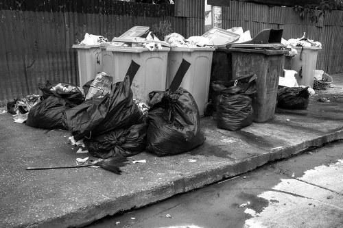 A row of four large wheelie bins with closed lids, positioned on a concrete pavement in an outdoor area, with the bins containing various types of waste. In front of the bins, there are several black refuse bags, some partially torn open, holding additional rubbish. The waste includes crumpled paper, plastic packaging, and miscellaneous clutter. To the right, a smaller black bin with a closed lid is visible, while in the background, a wooden fence and a garden space can be seen. The scene appears to be in a residential or commercial area, typical of rubbish storage before collection, and is associated with waste management services provided by Commercial Waste Removal Southfields, serving the Southfields area in London, SW19.