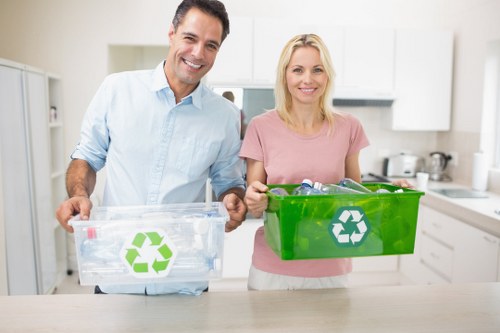 A smiling man and woman standing in a modern, bright kitchen, each holding a reusable recycling bin with a prominent green recycling symbol. The man on the left is wearing a light blue button-down shirt and is holding a clear plastic bin filled with recyclable bottles. The woman on the right is dressed in a soft pink short-sleeved top and is holding an opaque green bin. The kitchen features white cabinetry, stainless steel appliances, and a light countertop, creating a clean, organized environment. The scene conveys an emphasis on waste segregation and environmentally conscious disposal, relevant to rubbish removal services such as those offered by Commercial Waste Removal Southfields in the Southfields area, serving local recycling and waste collection needs.