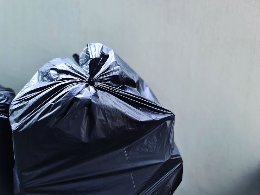 A person is seen disposing of paper waste into a grey rubbish bin, with their hand holding a crumpled brown paper bag that is partially inside the bin. The bin appears to be placed on a wooden surface
